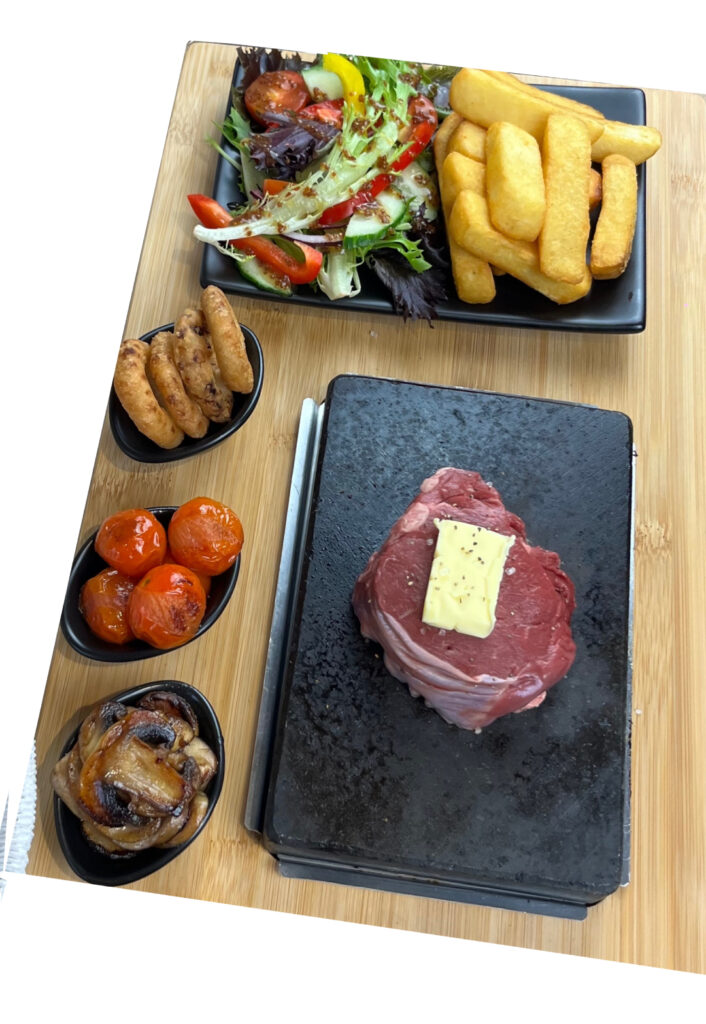 A mouth-watering meal featuring a fillet steak on a hot lava stone, with sides of grilled cherry tomatoes, sautéed mushrooms, and onion rings. A fresh mixed salad with chunky chips and a sprinkle chefs secret seasoning, make this a super sizzle night.
